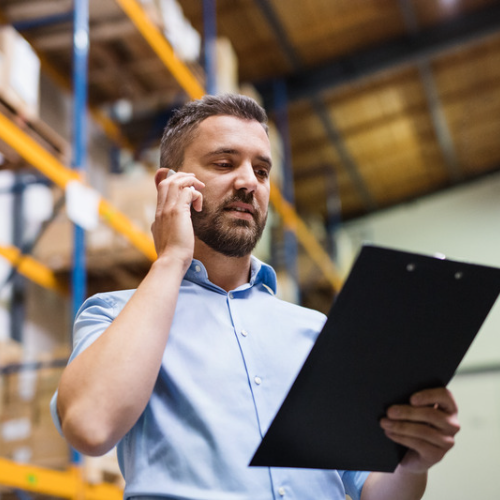 Caucasian male safety foreman making a phone call while looking at a clipboard in a warehouse facility