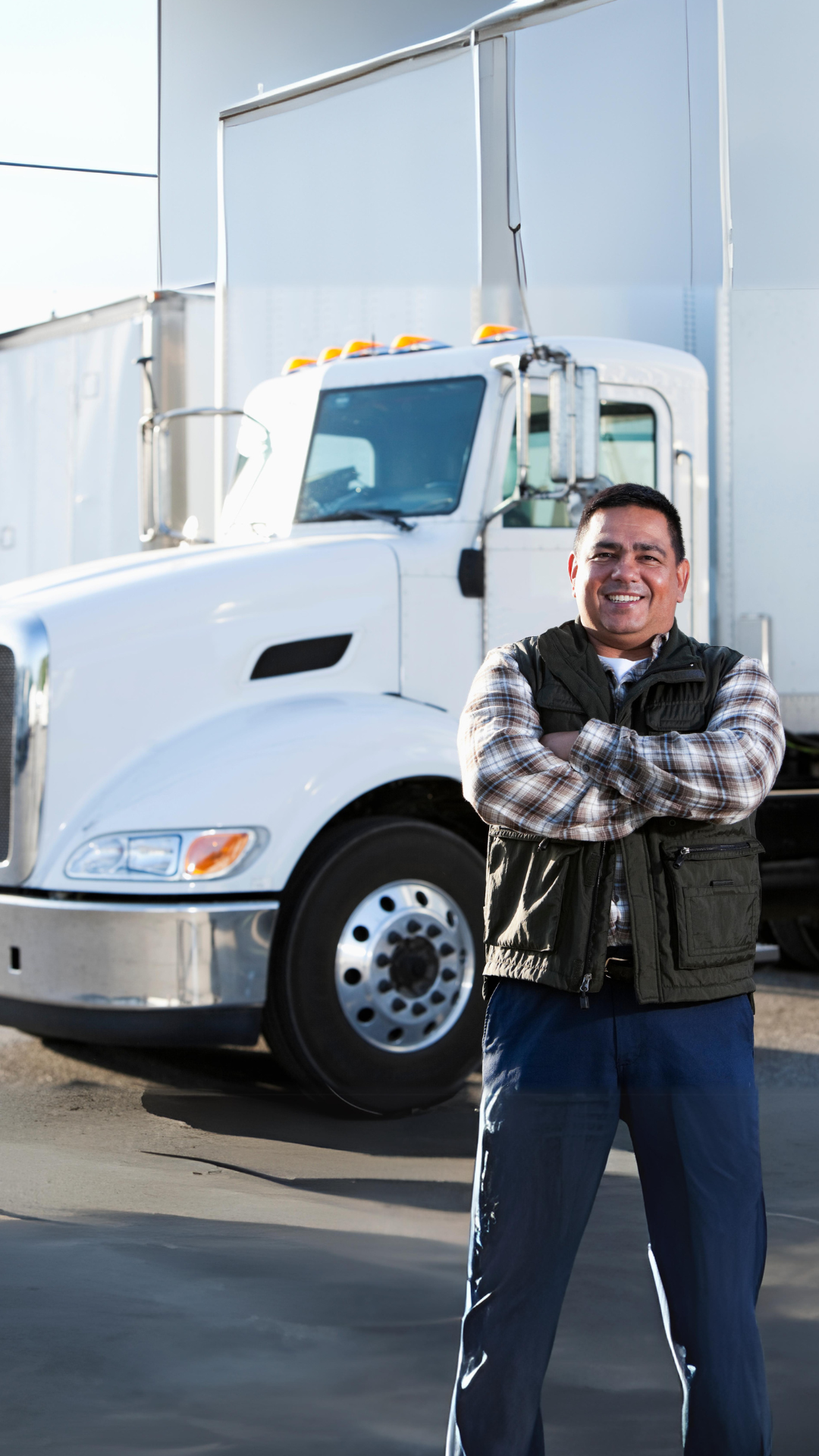 Latino male truck driver standing with arms crossed in front of white freight truck.