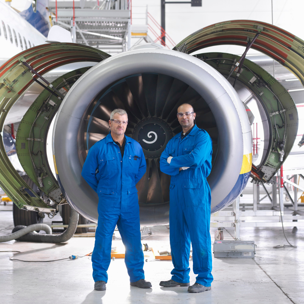 Two aerospace company employees wearing blue jumpsuits and standing in front of a turbine engine inside of a hanger 